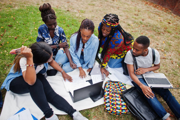 Group of five african college students spending time together on campus at university yard. Black afro friends sitting on grass and studying with laptops.